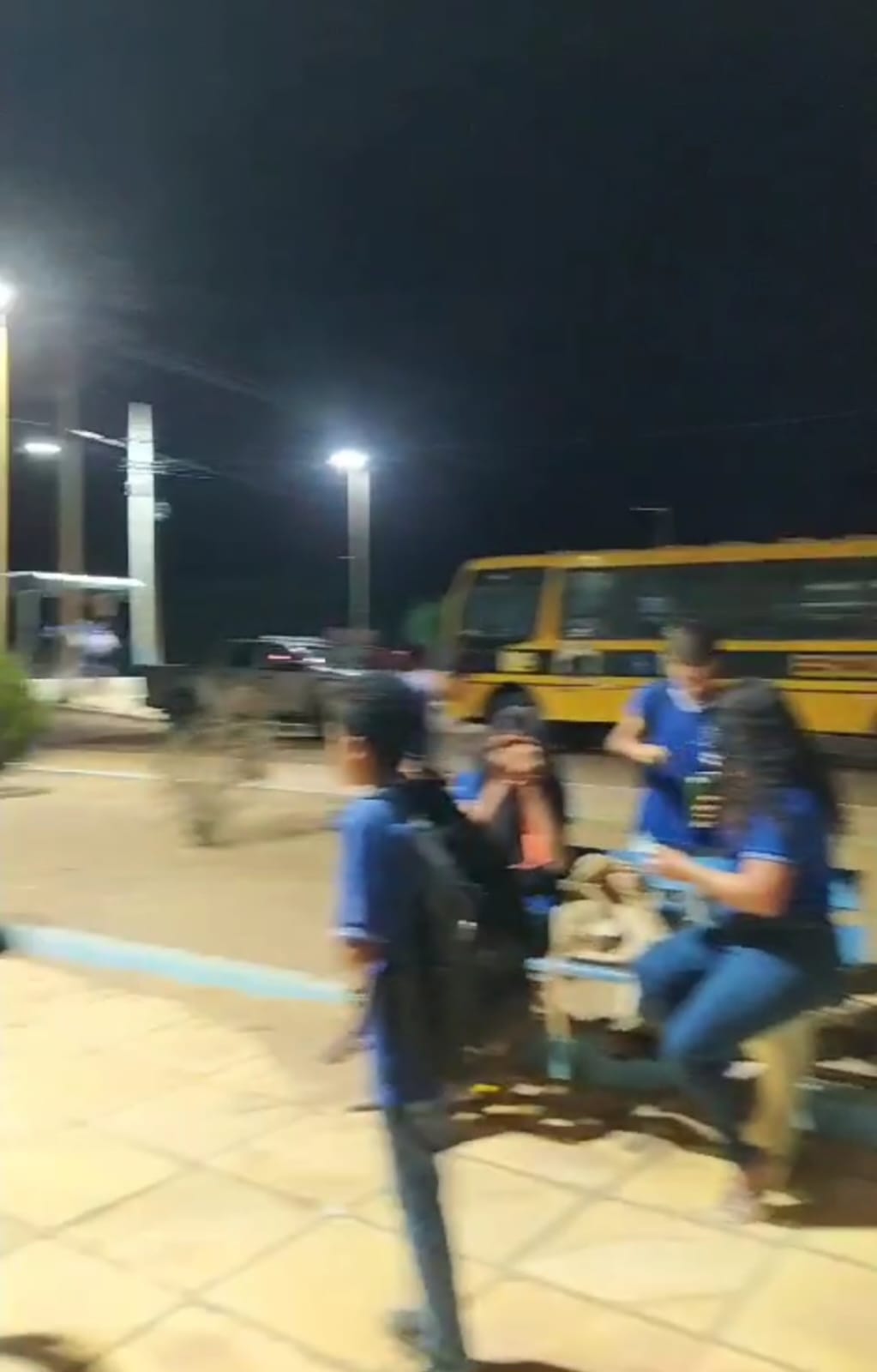 Estudantes da Escola Érico Veríssimo aguardam sentados na escola, enquanto o ônibus quebrado, ao fundo, ilustra a falta de transporte escolar na Serra dos Cláudios, Simoes, PI.